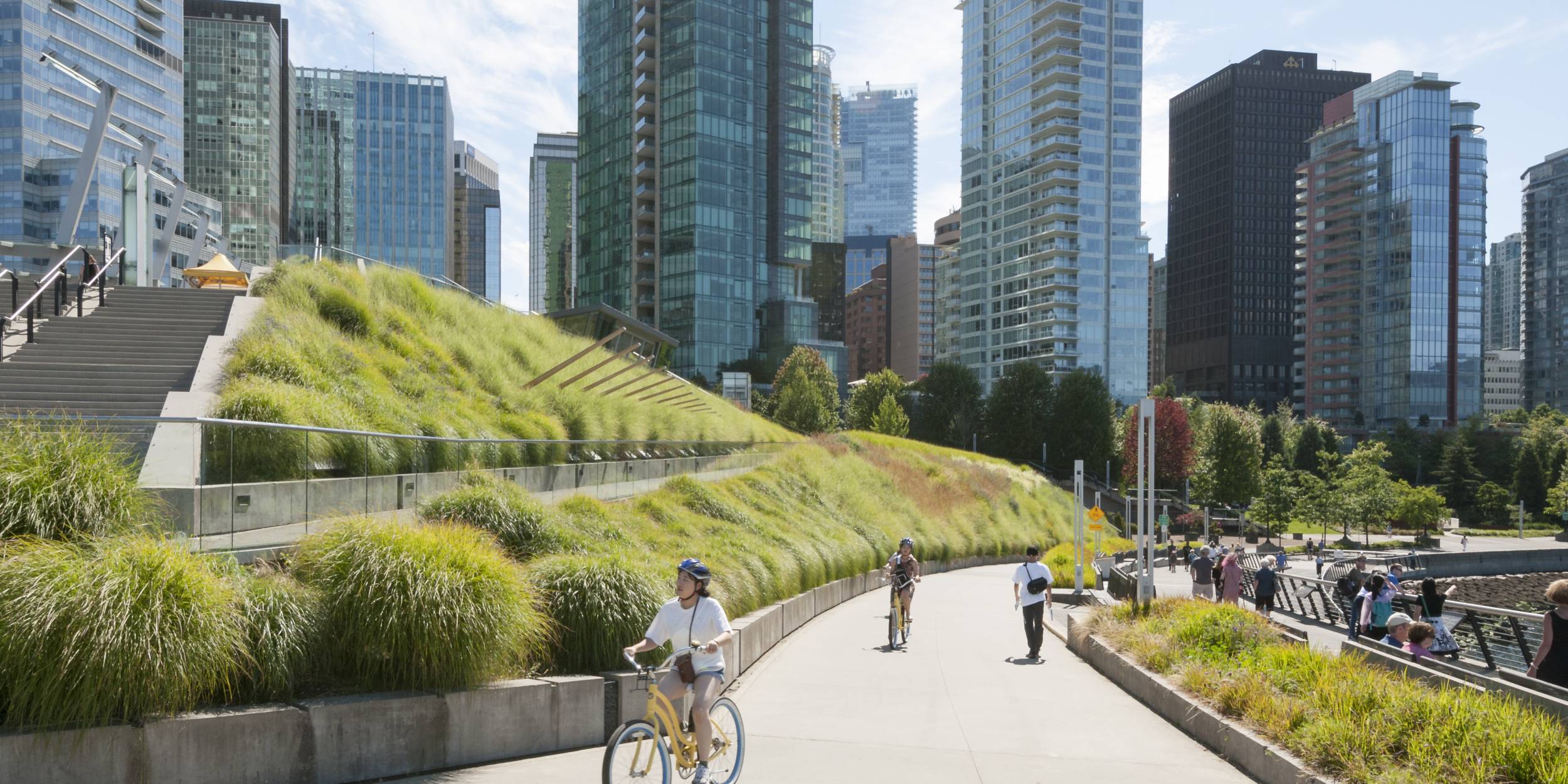 Photo of the Vancouver Convention Centre walkway where a person rides their bike in front of the lush green roof planting and pedestrians walk in the distance.