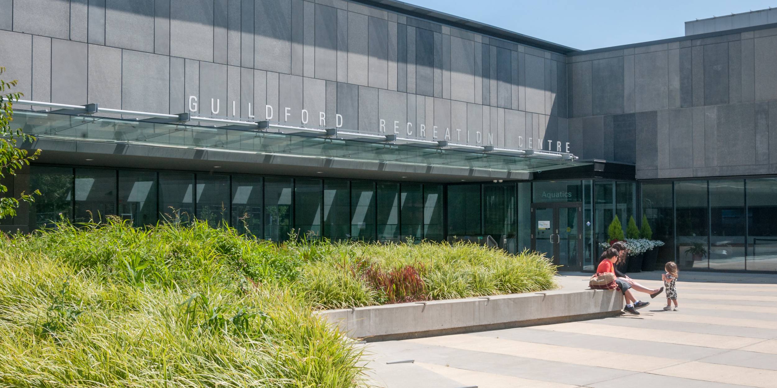 A family sits outside Guilford Aquatic Centre next to a large planting bed.