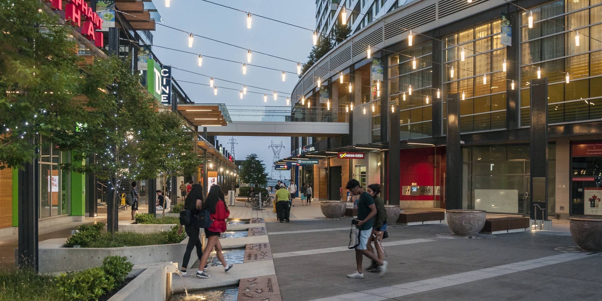 People funnel through Marine Gateway plaza as string lights hang above.
