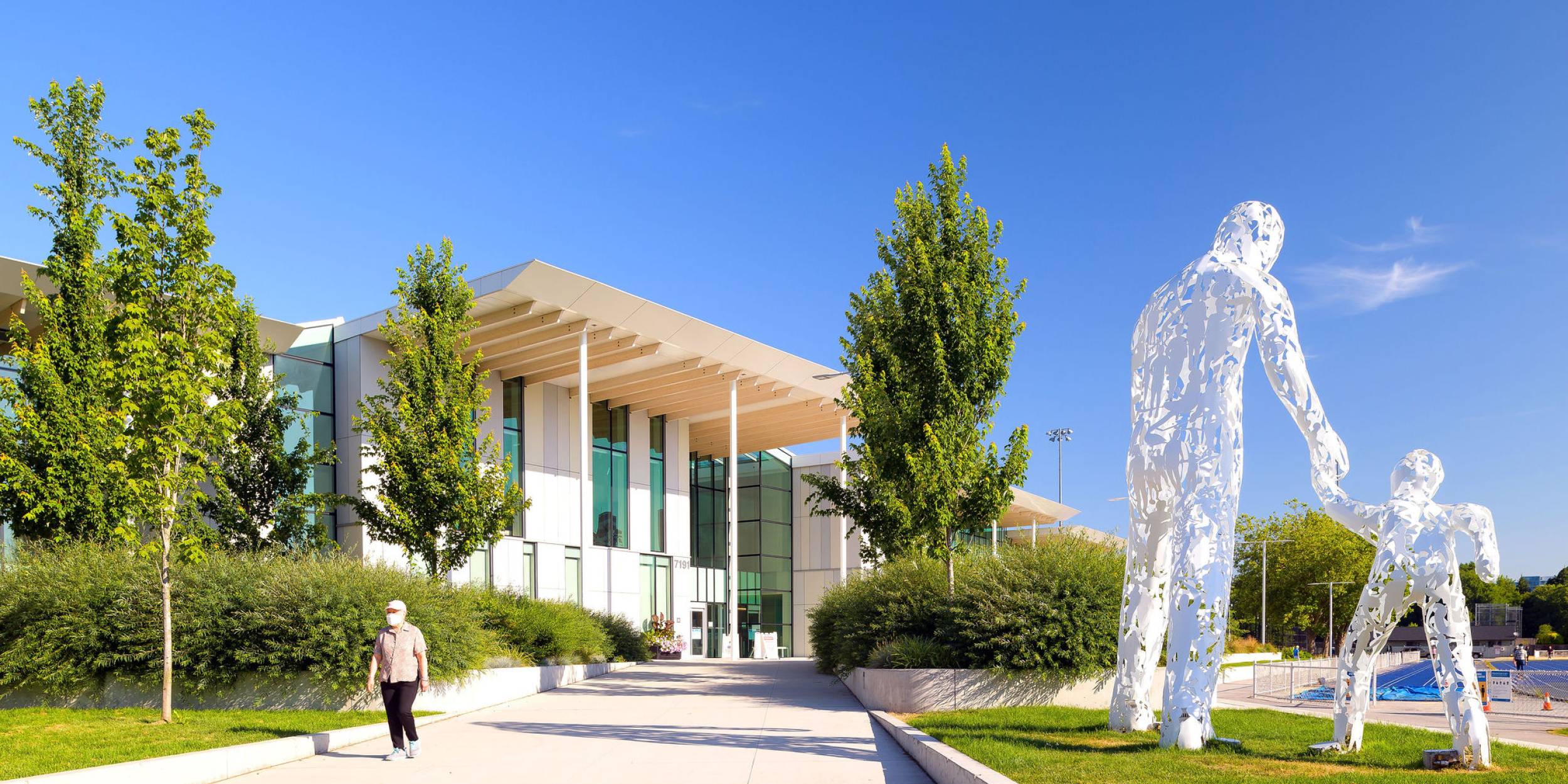 A man walks along the pathway to Minoru Aquatic Centre, adjacent to a large public art piece of an adult and a child holding hands.